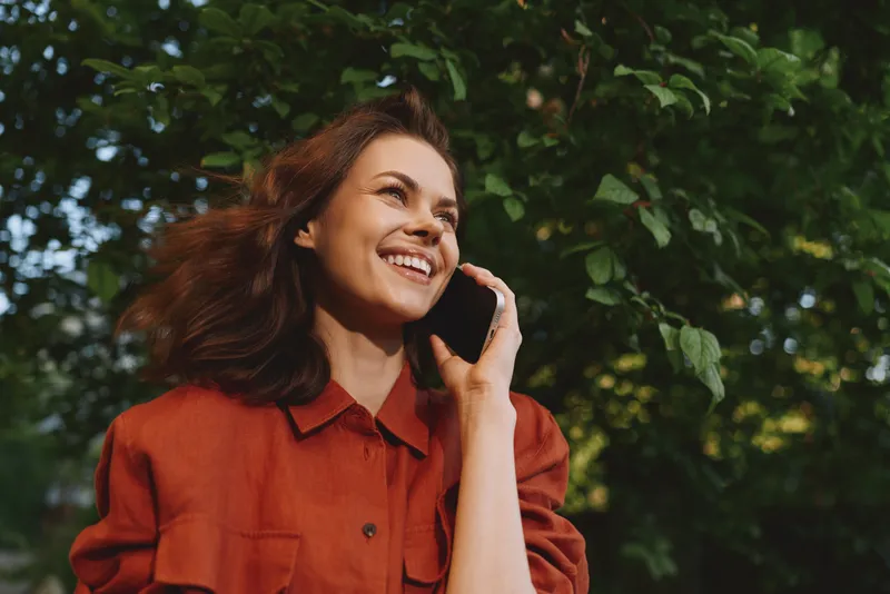 A smiling woman on a phone call outdoors in a bright orange jacket, framed by lush greenery. A candid lifestyle moment highlighting casual communication