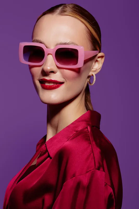 Stylish woman with pink sunglasses and red lipstick poses confidently against purple background. Fashion concept with glamour, elegance, and modern accessories in studio lighting.