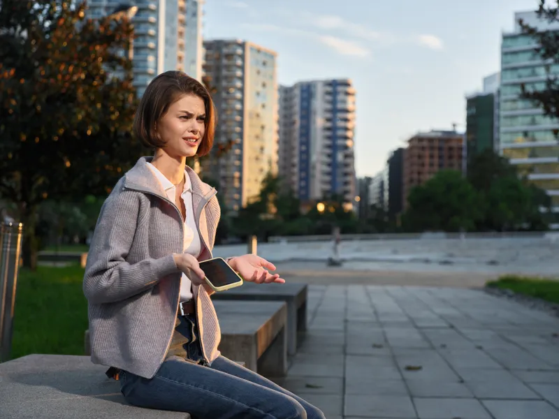 An urban woman sits on a bench in a modern park, texting on a smartphone. She wears a light jacket and jeans, smiling as she connects.