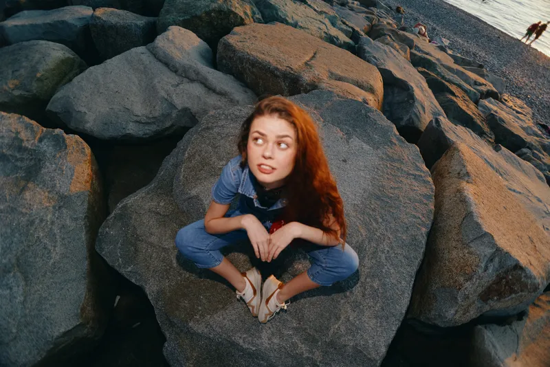 young woman sitting on large rocks wearing casual blue outfit sandals with red hair looking sideways in outdoor rocky environment during daylight