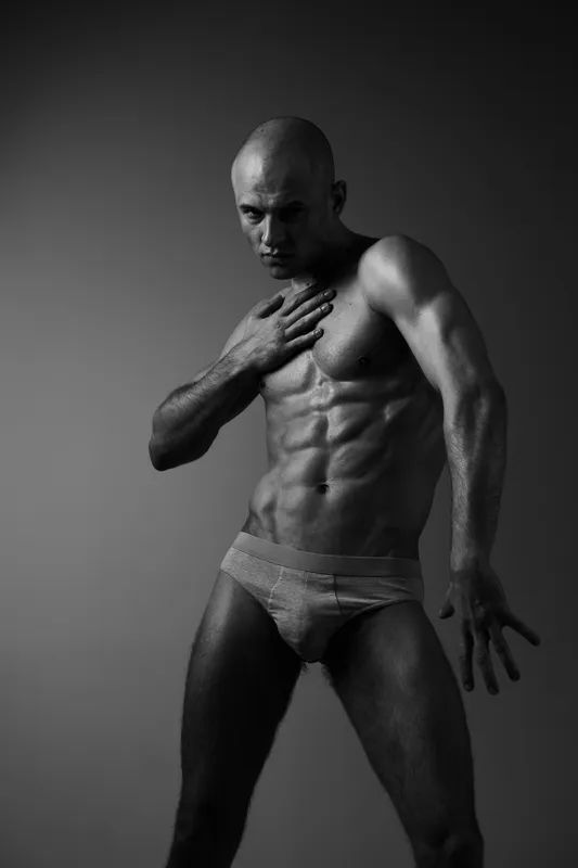 Black and white image of a muscular man posing in a dynamic stance, showcasing strength and masculinity with a dramatic silhouette.