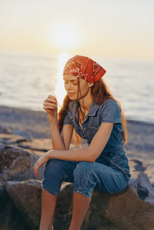 Lifestyle woman in modern boho-western denim street style wearing a red bandana, sitting on rocks by the sea at sunset, film color warm tones capturing natural mood.