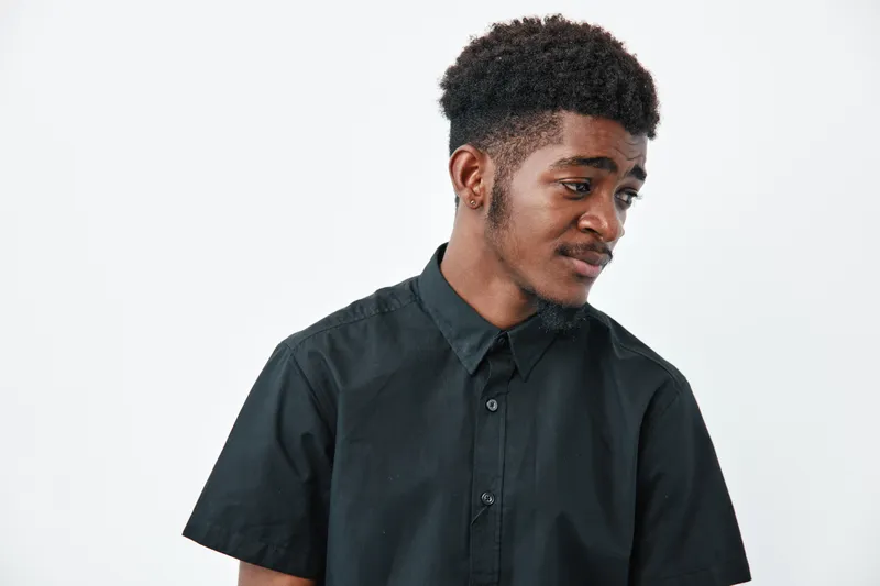 Young African American man with dark skin and curly hair wearing black shirt looking sideways on white background. Portrait, casual style and thoughtful expression in studio.