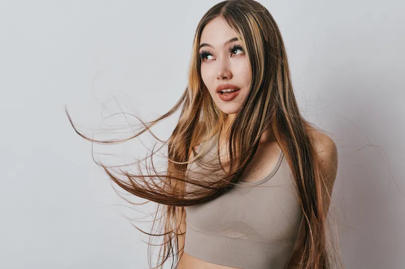 Young woman with long hair blowing and wearing tank top looking to side with open mouth on neutral background. Portrait of female model with casual style and natural beauty.