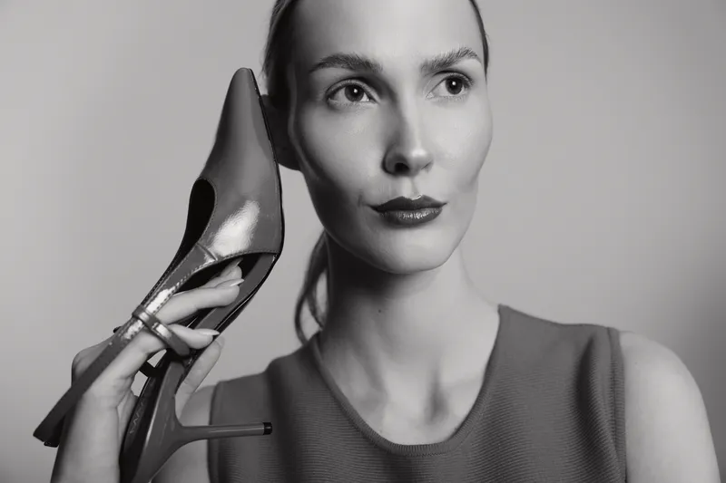 Portrait of a serious Caucasian woman with pale skin holding a high heel shoe near her face on a neutral background.
