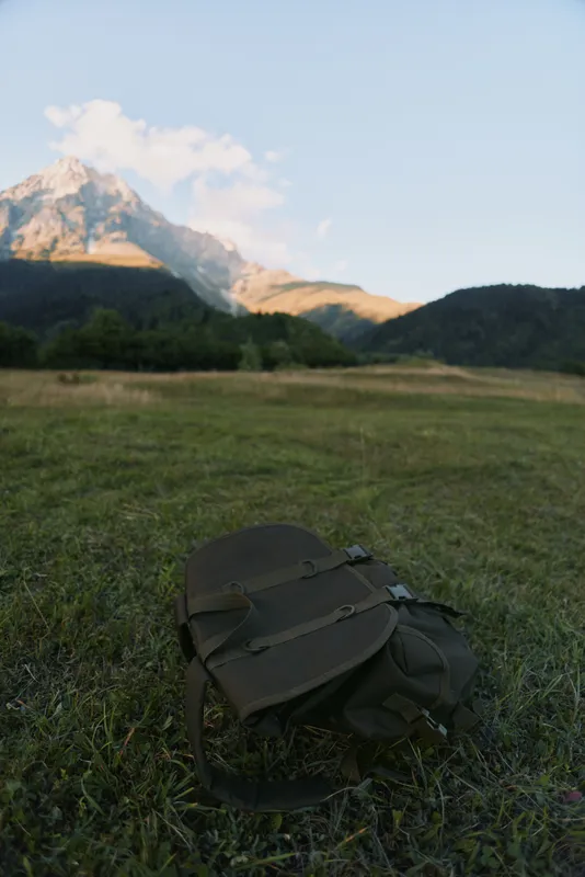 A green backpack lies on a grassy field with a mountain peak illuminated by sunlight in the background under a clear blue sky.