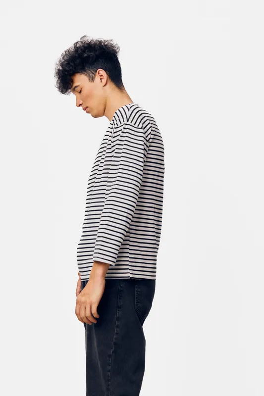 Young man wearing a striped long sleeve shirt stands sideways with a neutral expression against a white background, showcasing casual youth fashion in soft lighting.