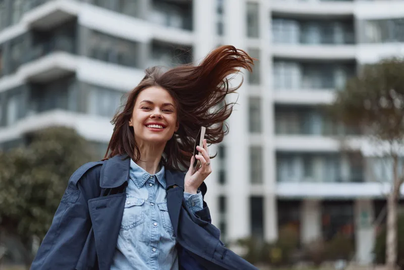 Young woman wearing a denim shirt and navy coat walks along a city street while talking on her phone, smiling with wind in her hair and a casual confident mood.