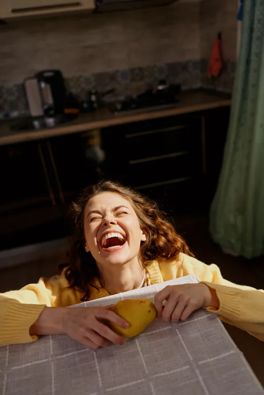 Young woman with curly hair laughs while gripping a bright yellow lemon over a table in a cozy home kitchen.