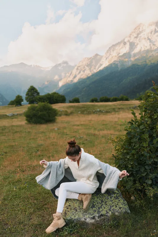 Woman outdoors in a meadow near mountains, wearing a light sweater and white pants, sitting on a rock with a cozy cape, enjoying a relaxed sunlit moment.