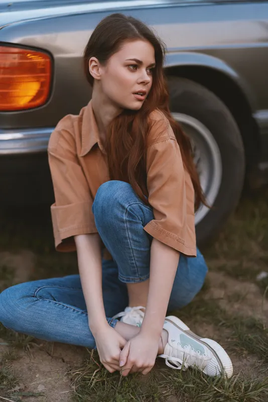 Young woman sitting casually by a vintage car, wearing stylish denim and a brown shirt, expressing a relaxed emotion amidst a natural background.