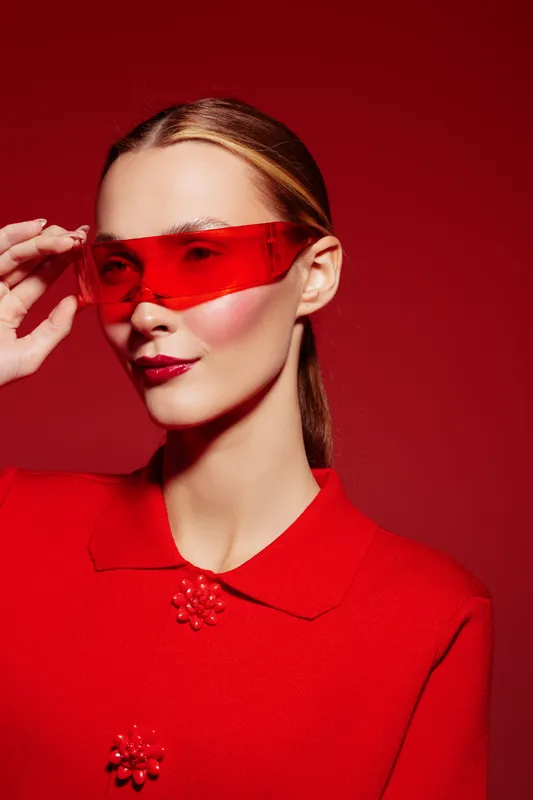 Stylish woman in red dress and red translucent glasses poses against red background. Fashion portrait with sleek hairstyle and bold makeup. Concept of modern style and confidence.