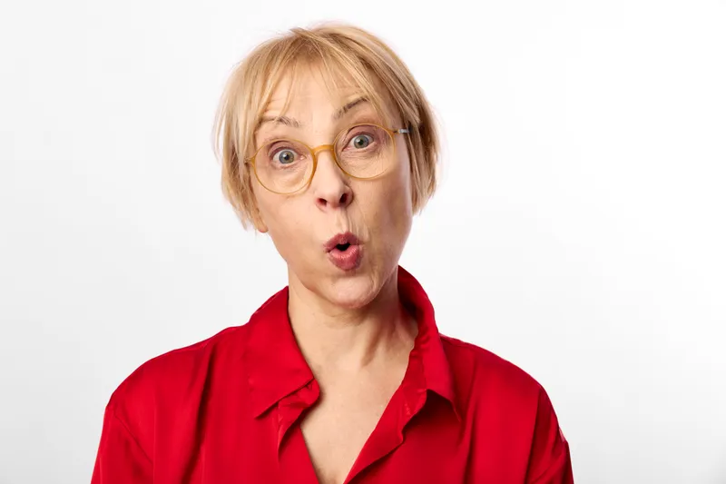 Woman surprised with glasses, red shirt, close up portrait on white background. Face expression with open mouth conveys shock and emotion in soft studio light calm wellness mood.