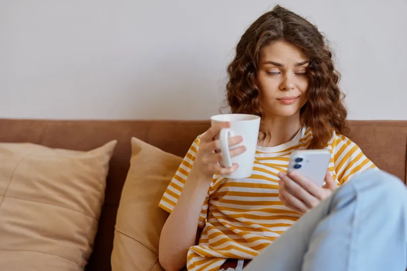 A woman relaxing at home, sitting on a sofa, holding a mug and checking her smartphone, showcasing casual lifestyle and indoor comfort in a cozy living space.