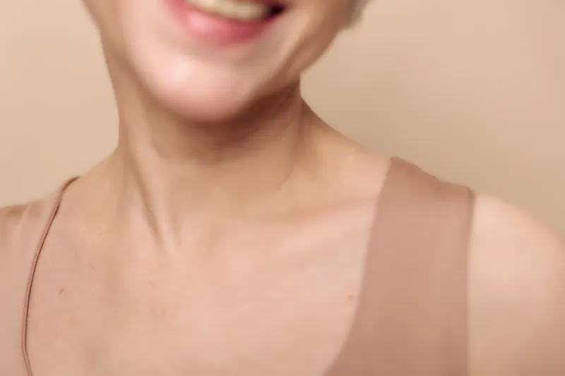 Closeup of smiling woman with natural skin tone wearing beige top, showing neck and shoulders. Concept of beauty, skin health, and positive mood in soft lighting.