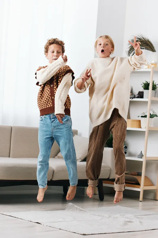 Two joyful children jump mid air indoors wearing cozy knit sweaters and jeans in a bright living room. A candid, playful moment with warm, cheerful atmosphere.