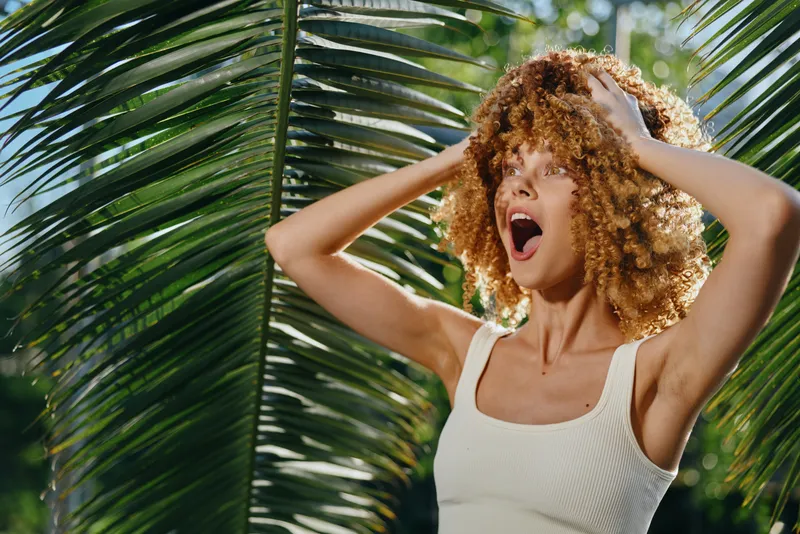 surprised woman with curly hair standing near large tropical leaves in bright sunlight, summer nature background with vibrant green palm fronds