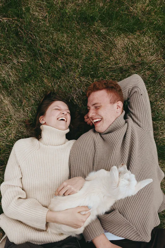 Joyful couple lying on grass with a fluffy white cat, warm sweaters, relaxed mood, outdoor lifestyle moment, natural light, candid scene, gentle smiles, cozy textures, peaceful atmosphere