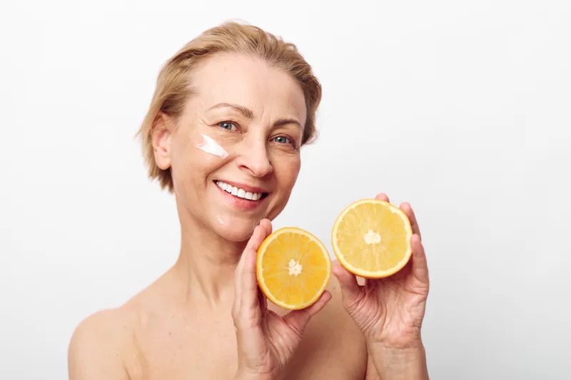 Smiling middle aged woman with natural skin and facial cream holds two halves of fresh orange. Bright studio lighting on white background. Concept for skincare, health, and natural beauty care.