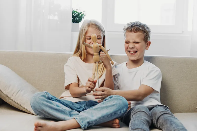 Two children sitting on sofa playing with wooden mannequin, smiling and enjoying playful moment in bright living room with natural light.