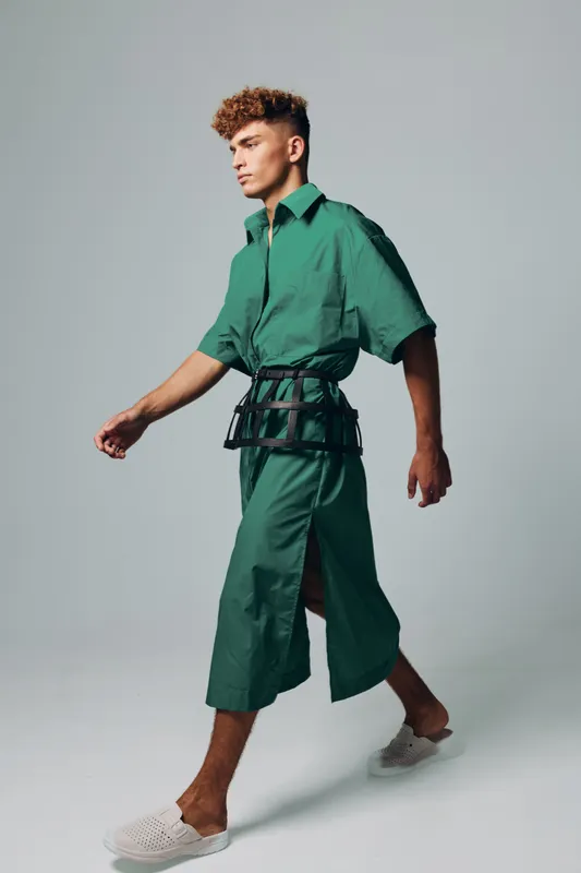 Young man walking in green oversized shirt and wide cropped pants with black belt accessory and white slip on shoes on gray background in studio.