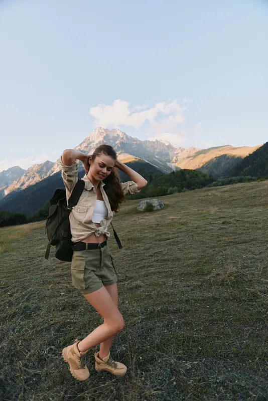 Young woman with long hair enjoys a mountain hike wearing a beige shirt tied at the waist and green shorts with hiking boots in a scenic outdoor setting.