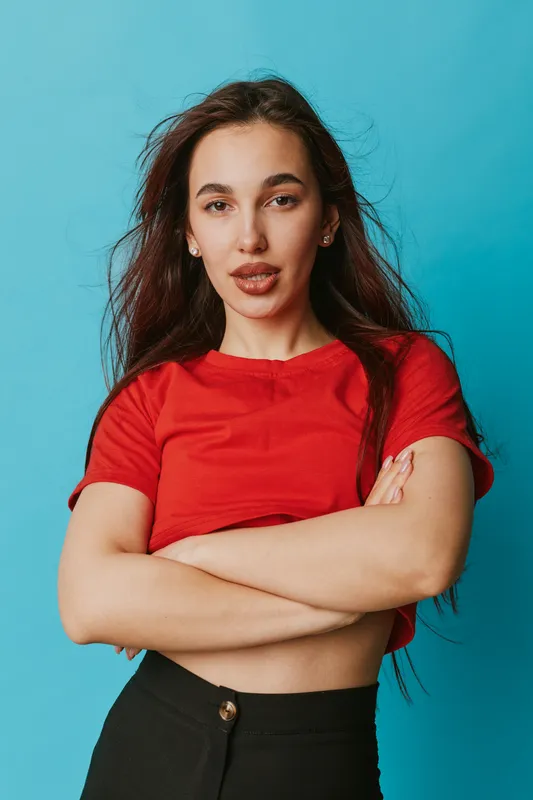 Young woman with long hair wearing red shirt and black pants standing with crossed arms against blue background looking confident and relaxed in studio portrait.