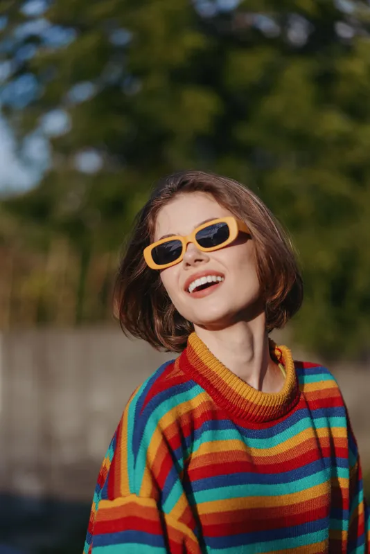 Woman young adult smiling in a colorful striped sweater and yellow sunglasses, casual outfit portrait outdoors in warm sunlight, bob haircut, looking up, joyful lifestyle and fashion image