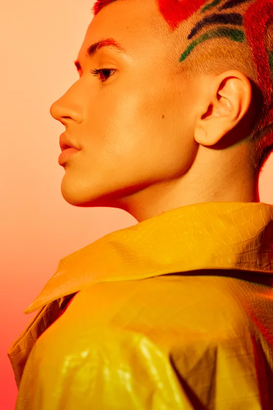 young woman with stylish short hair and colorful shaved patterns wearing a yellow jacket, glowing skin and warm orange background striking a modern urban look