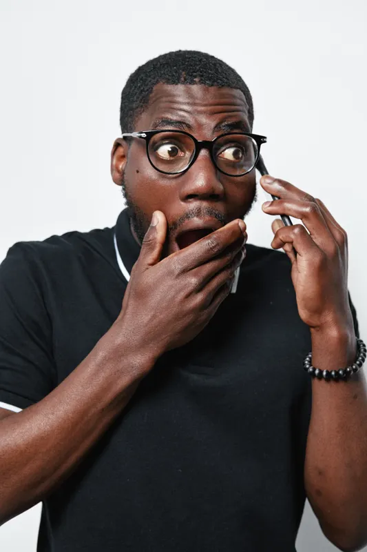 African American man with dark skin and glasses talking on smartphone with surprised expression covering mouth with hand in casual black polo shirt on white background.