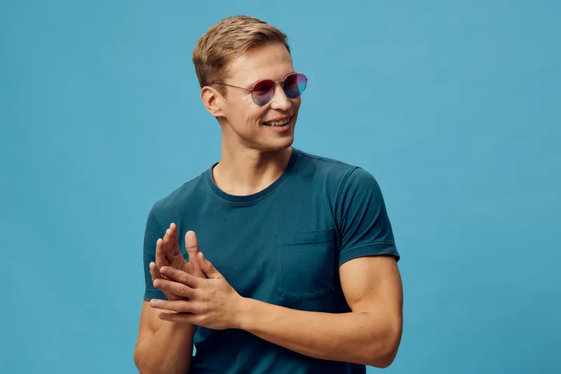 Smiling young man with sunglasses wearing casual teal t-shirt against bright turquoise background, expressing happiness and friendliness, studio portrait for lifestyle and fashion concepts.