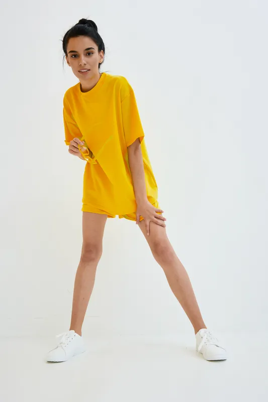 Young woman in bright yellow oversized t-shirt posing confidently against white background, wearing white sneakers and casual hairstyle, modern minimal style.