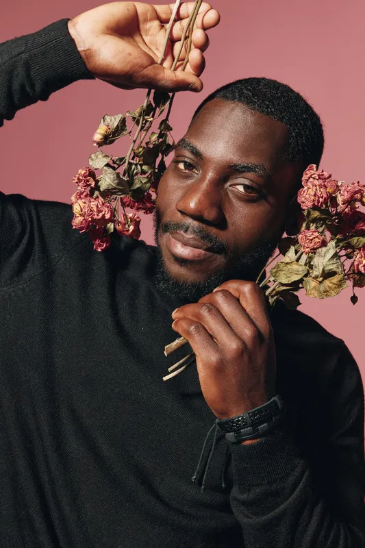 Creative conceptual portrait of African man holding dried flowers with thoughtful expression in visual style. Dark skin, black sweater, and pink background emphasize artistic mood and culture.
