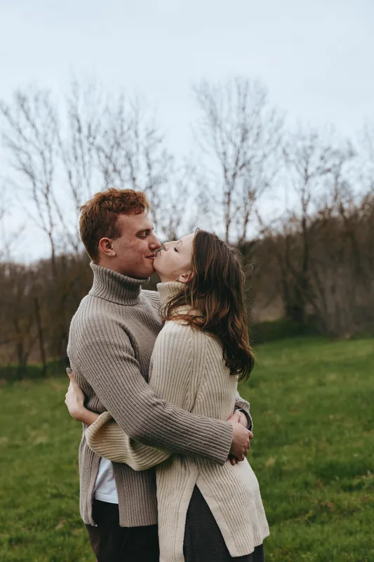 Two people share a warm kiss and embrace in a grassy park, wearing cozy sweaters, captured in natural daylight. A candid moment conveying romance, affection, and lifestyle, with calm outdoor ambiance