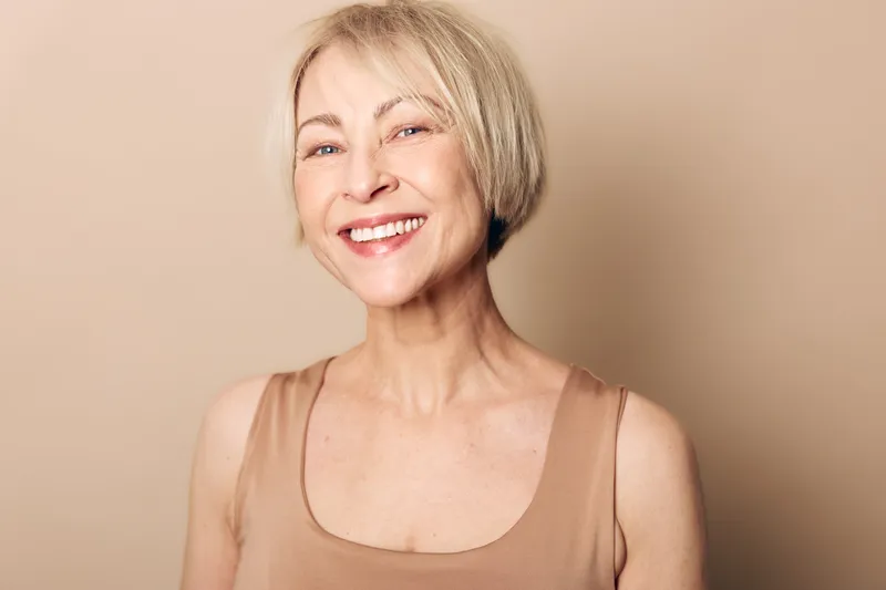 Smiling mature woman with short blonde hair wearing beige tank top stands against soft beige background. Concept of natural beauty, confidence, aging gracefully and wellness.