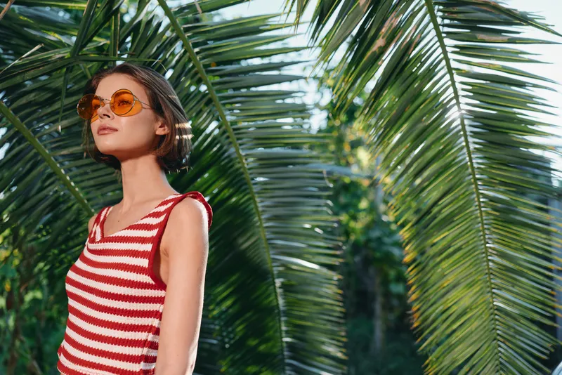A stylish woman stands among lush palm leaves, wearing a red and white striped tank top and round sunglasses. She exudes confidence and brightness in a sunny tropical setting.