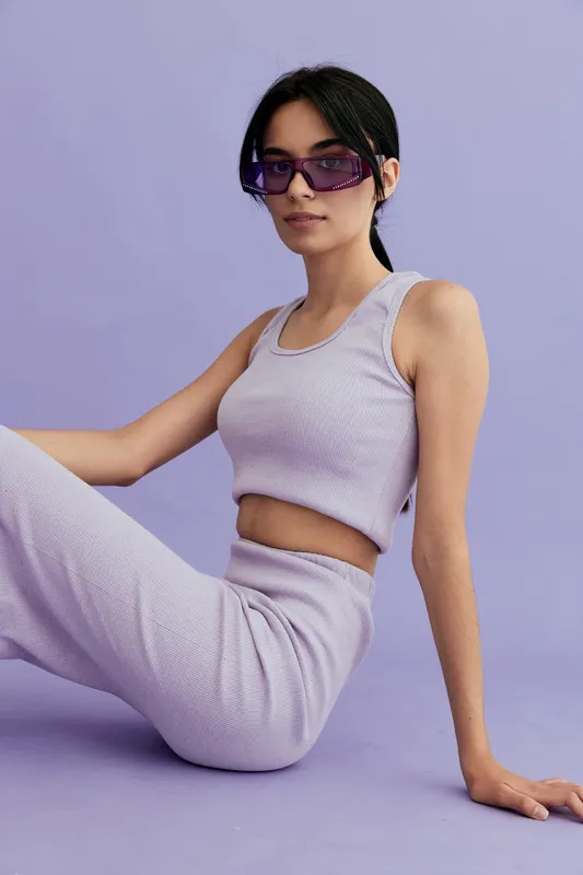 young woman sitting wearing lilac sleeveless top and matching pants with purple sunglasses against pastel purple background, casual relaxed pose and modern style
