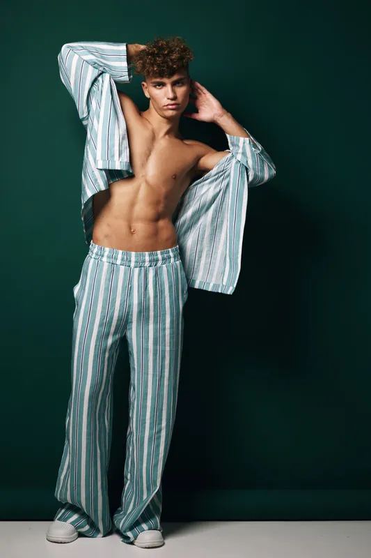 Young man posing in striped pajamas with open shirt showing toned abs, standing against dark background in studio. Fashion, casual wear and confident male model with curly hair.