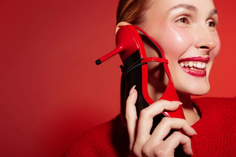 Smiling woman holds red high heel shoe near ear like phone, wearing red sweater on red background. Concept of fashion, fun, communication, and creative lifestyle portrait.