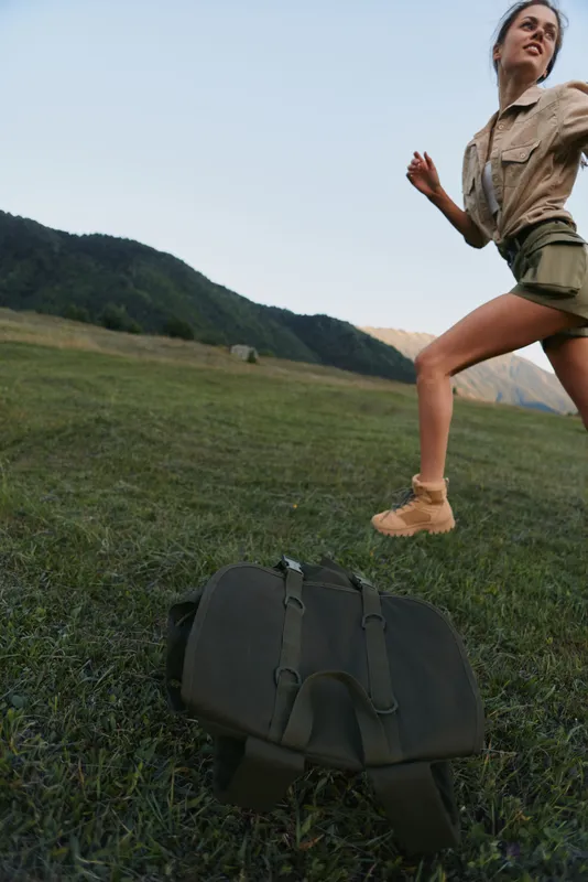A woman runs across a grassy field outdoors, a backpack secured at her side, capturing a dynamic moment of outdoor activity and adventurous lifestyle under bright blue sky.