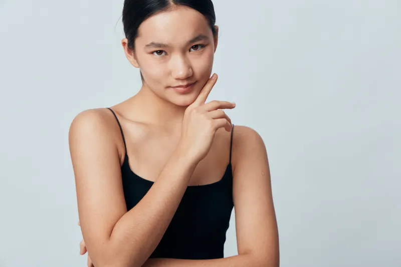 Young woman in black sleeveless top posing with hand near face, looking at camera with slight smile and confident expression against plain background in studio.
