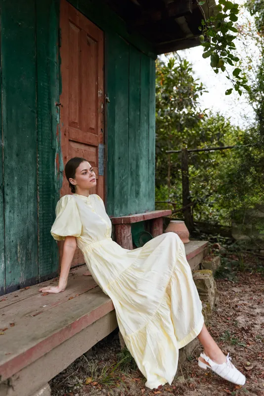 woman in a pale yellow dress sits on a wooden porch beside a green door, outdoor garden setting with rustic charm and a calm, stylish atmosphere