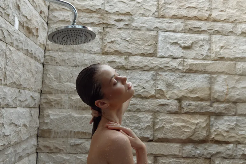 Relaxed woman enjoying warm shower with closed eyes under rain-style showerhead in beige stone bathroom, calm atmosphere, wellness and personal care concept.
