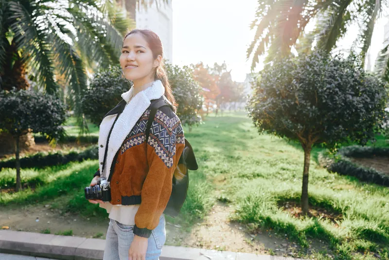 Woman with camera strolling through a sunny park, autumn light, green lawn and trees, backpack, casual sweater, candid outdoor photography scene.
