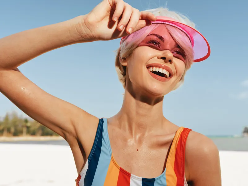 Bright beach scene featuring a smiling woman in a colorful striped swimsuit and pink visor, enjoying sun, breeze, and warm sand under a clear blue sky.