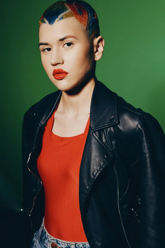 Young woman with colorful short hair wearing red top and black leather jacket standing confidently against green background with bold makeup and casual style.