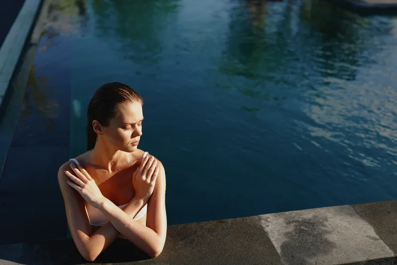 Young woman in swimming pool, relaxing with crossed arms, sunlight on skin, calm water background, peaceful moment, summer lifestyle, natural beauty