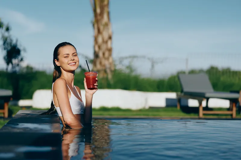 Young woman in a white swimsuit relaxes in an outdoor pool while holding a red drink and smiling with closed eyes on a sunny day.