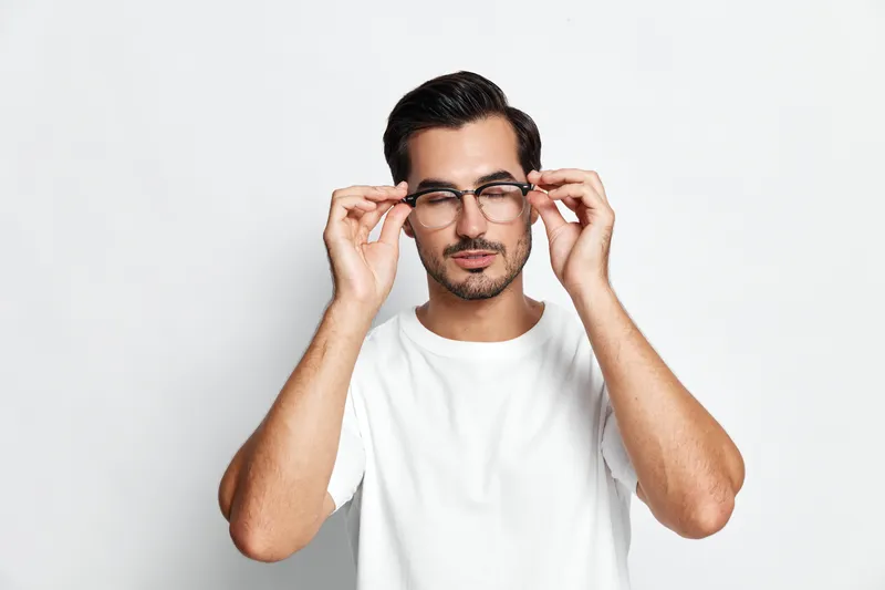 Confident man wearing glasses adjusting them with hands, dressed in casual white t-shirt, looking at the camera on solid light background. People lifestyle concept.