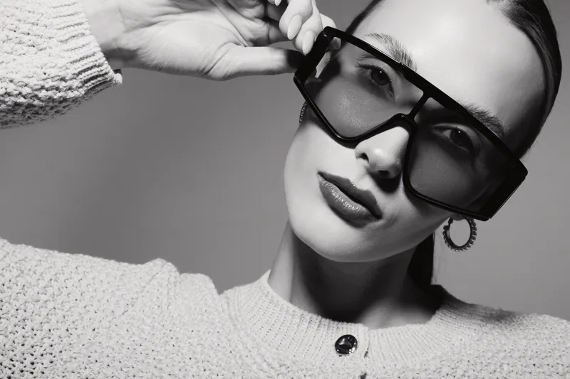Stylish white woman with light skin wearing large sunglasses and cozy sweater poses in studio with confident expression. Fashion and elegance concept.